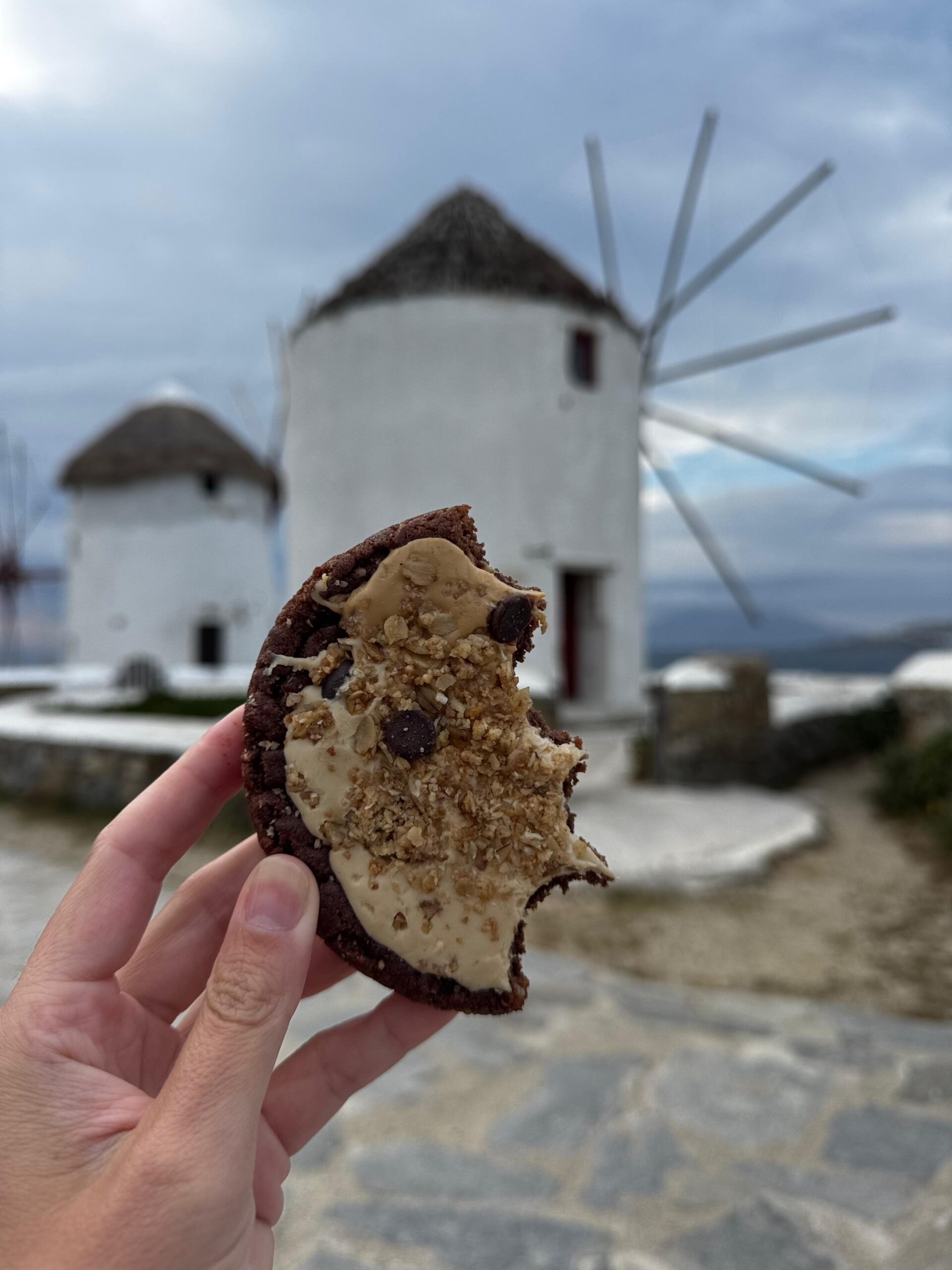Cookie com pasta e granola em frente aos moinhos de Mykonos, exemplo de lanche equilibrado em viagem